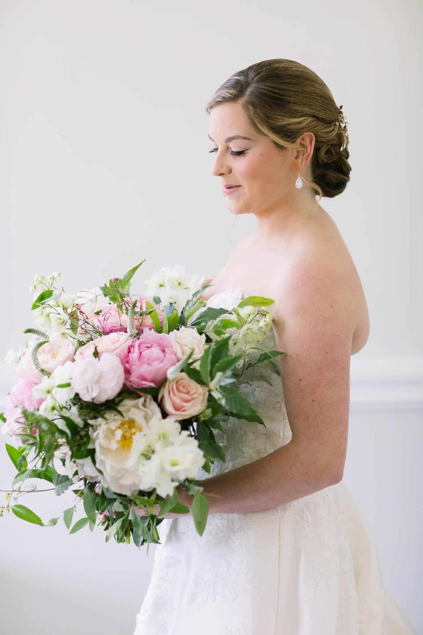 Bride in a strapless wedding gown holding a lush bouquet of pink peonies, roses, and white flowers, showcasing a joyful and elegant moment before her Boston wedding.