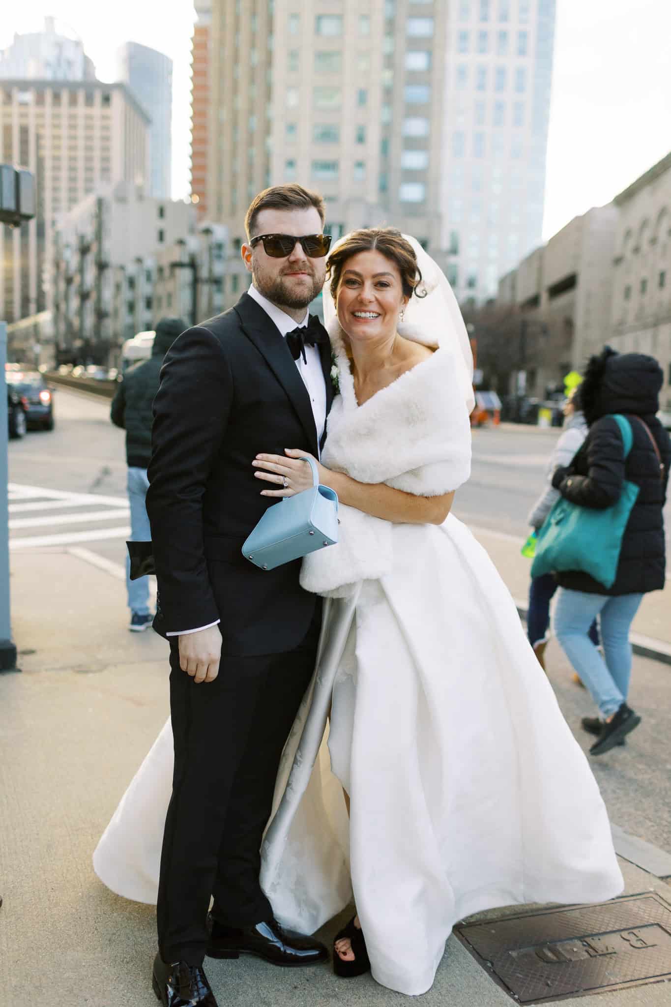 bride wearing a white fur shawl and groom in a tux pose outside their fairmont copley wedding planned by boston wedding planner Contagious Events